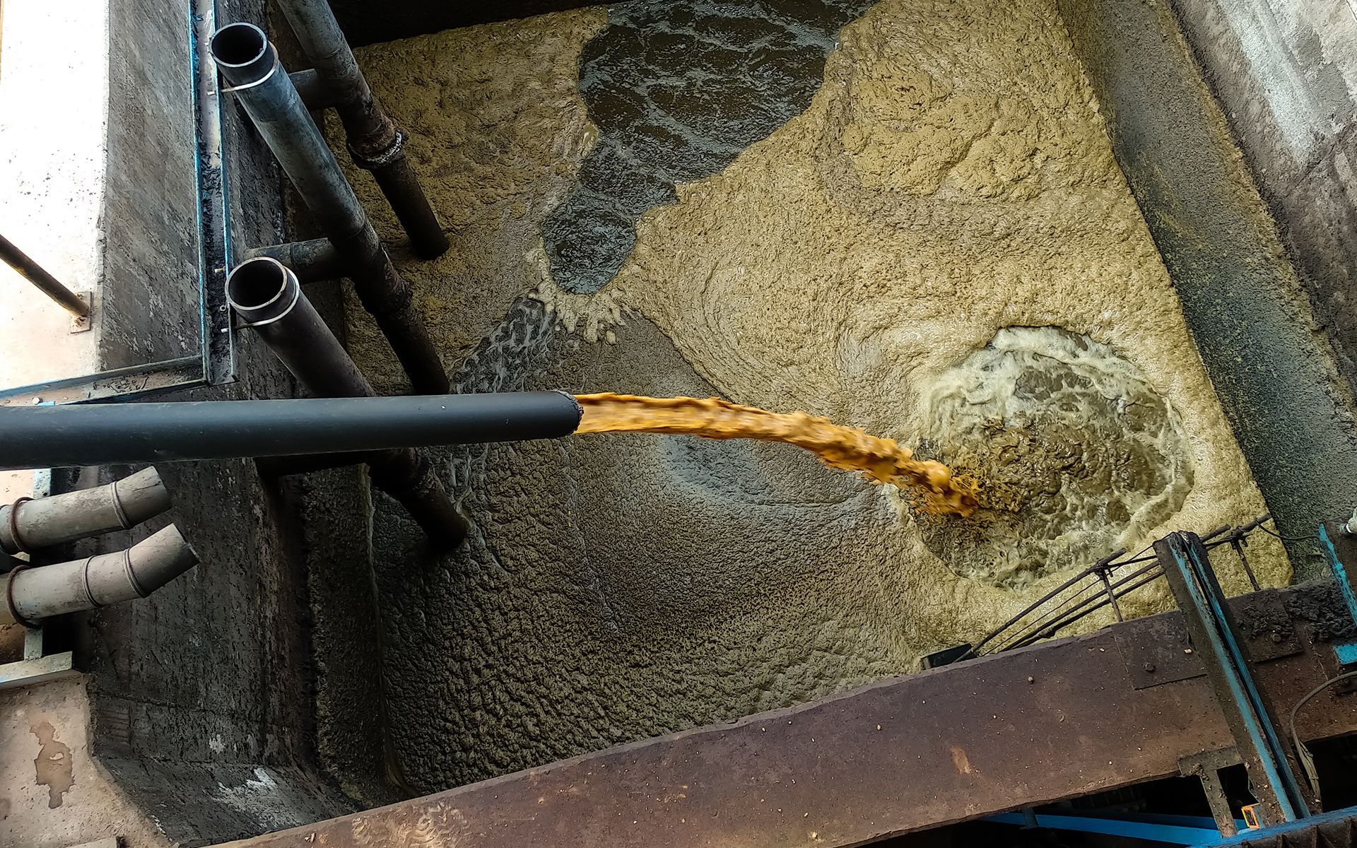 Brown liquid pouring into a large industrial tank filled with tan and gray sludge.