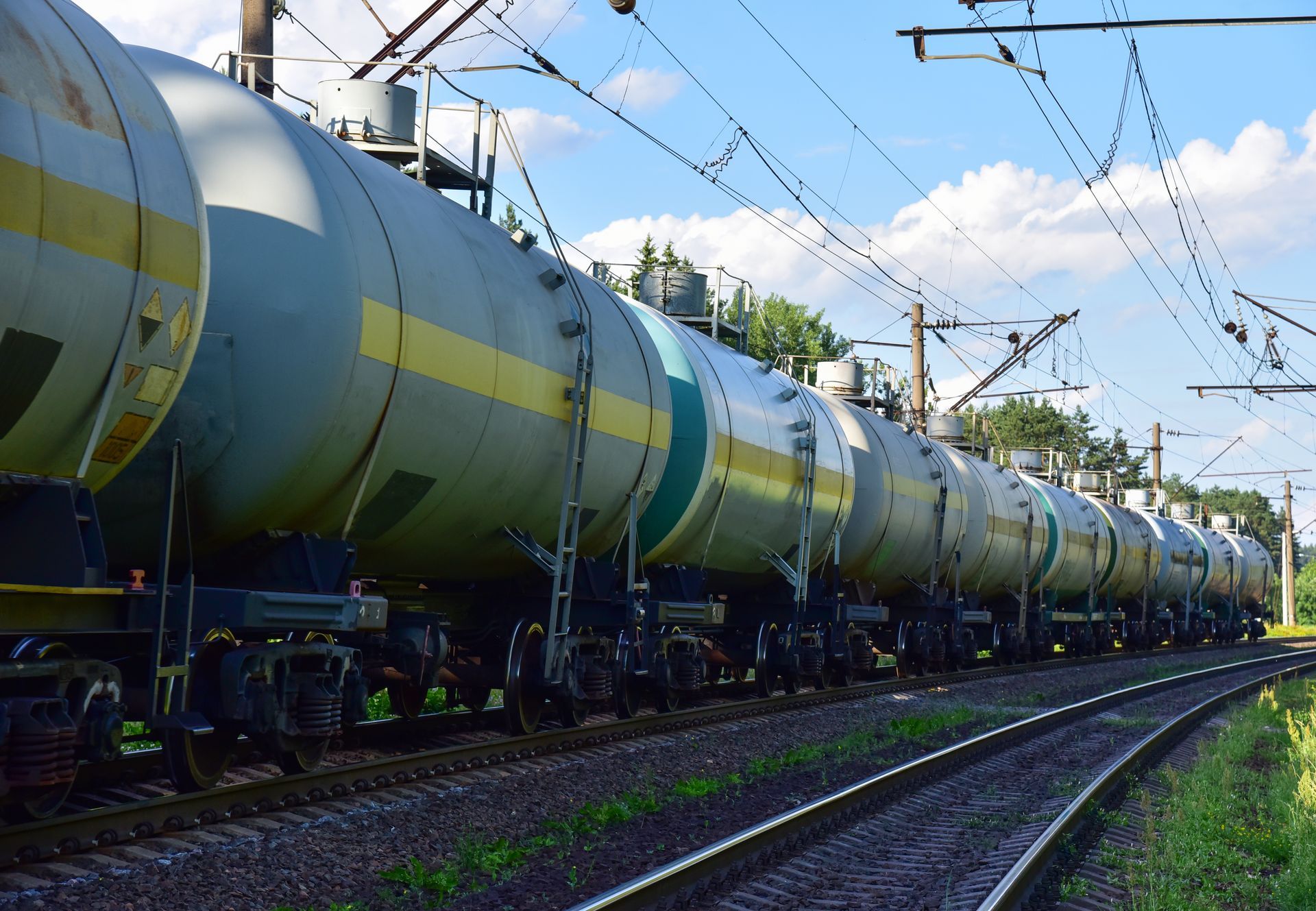 Tanker train cars on tracks curving right, with power lines above and greenery to the side.
