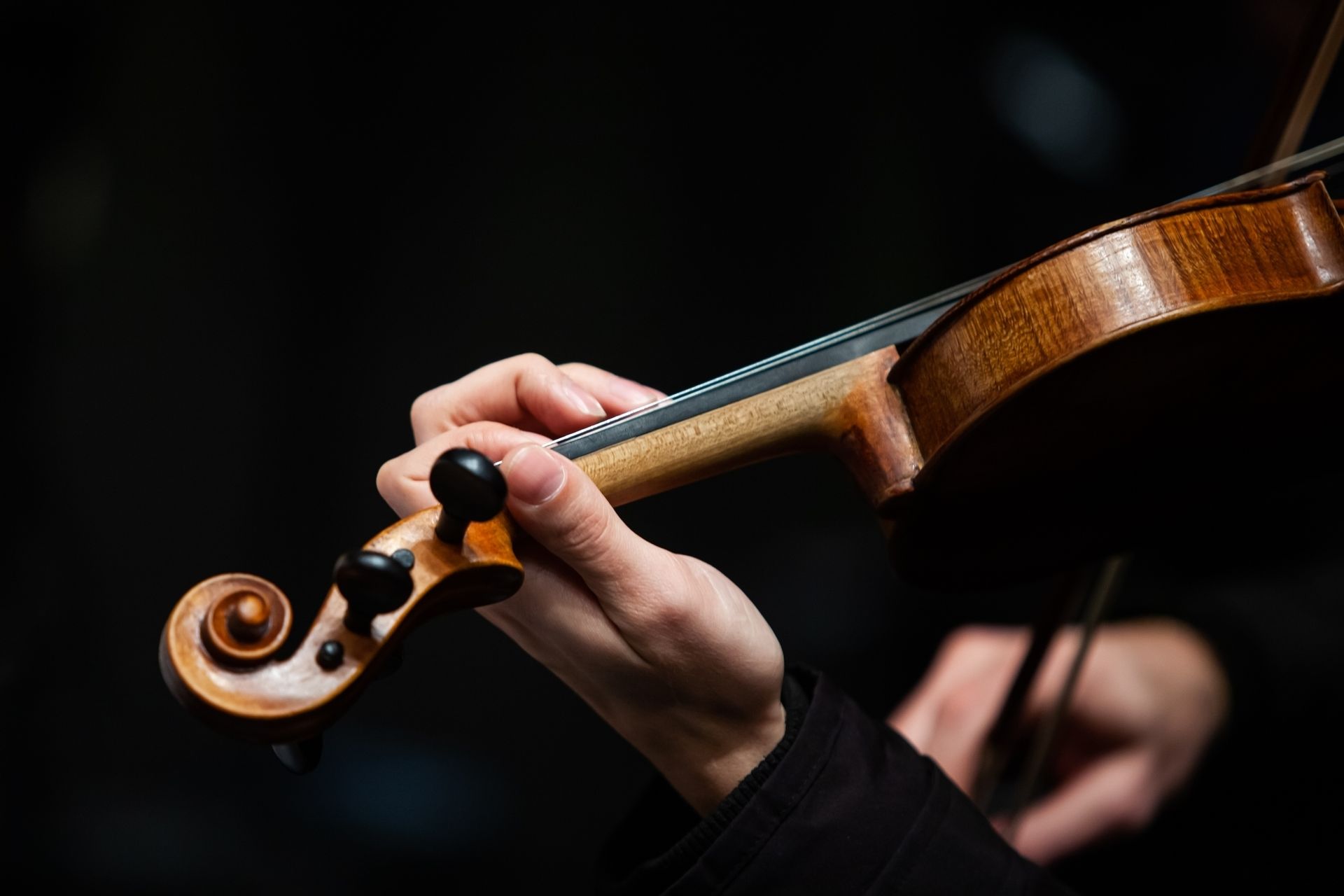 Close-up of a hand playing a violin with a bow; dark background.