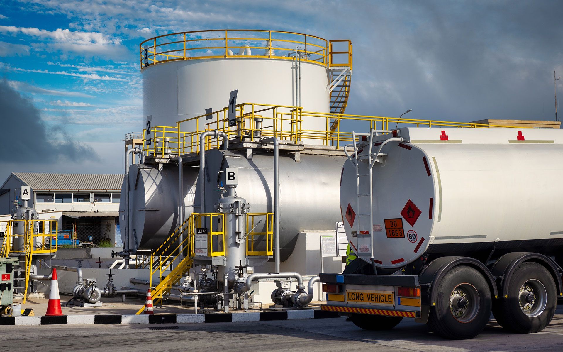 Tanker truck loading at fuel storage tank facility; overcast sky.