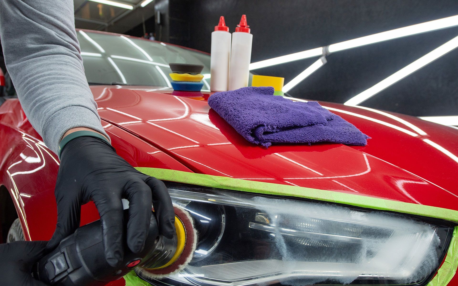 Person polishing a red car headlight with a power tool, surrounded by detailing supplies.