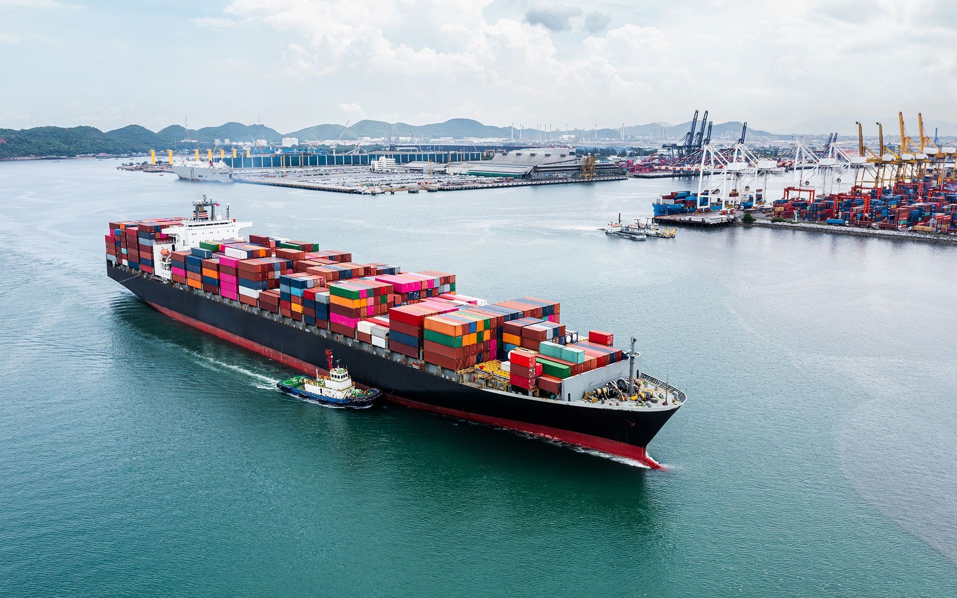Container ship sailing in harbor, loaded with cargo containers, assisted by a tugboat, port in the background.