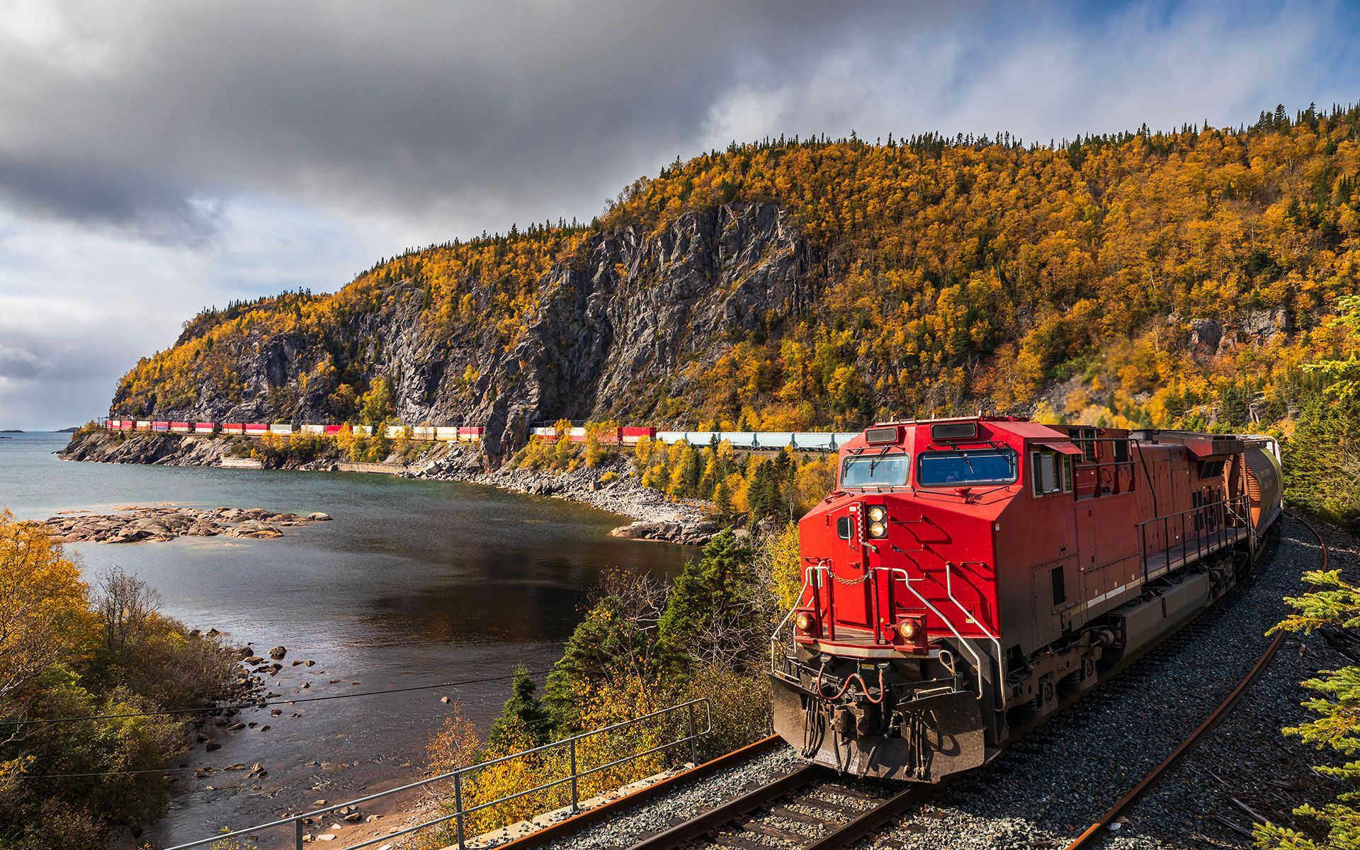 Red train travels along a coastal track during fall, beside a rocky shoreline and colorful hillside.