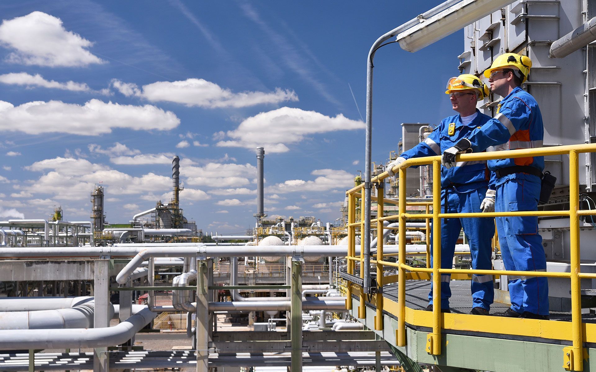 Two workers in blue coveralls and yellow helmets overlook an industrial plant.