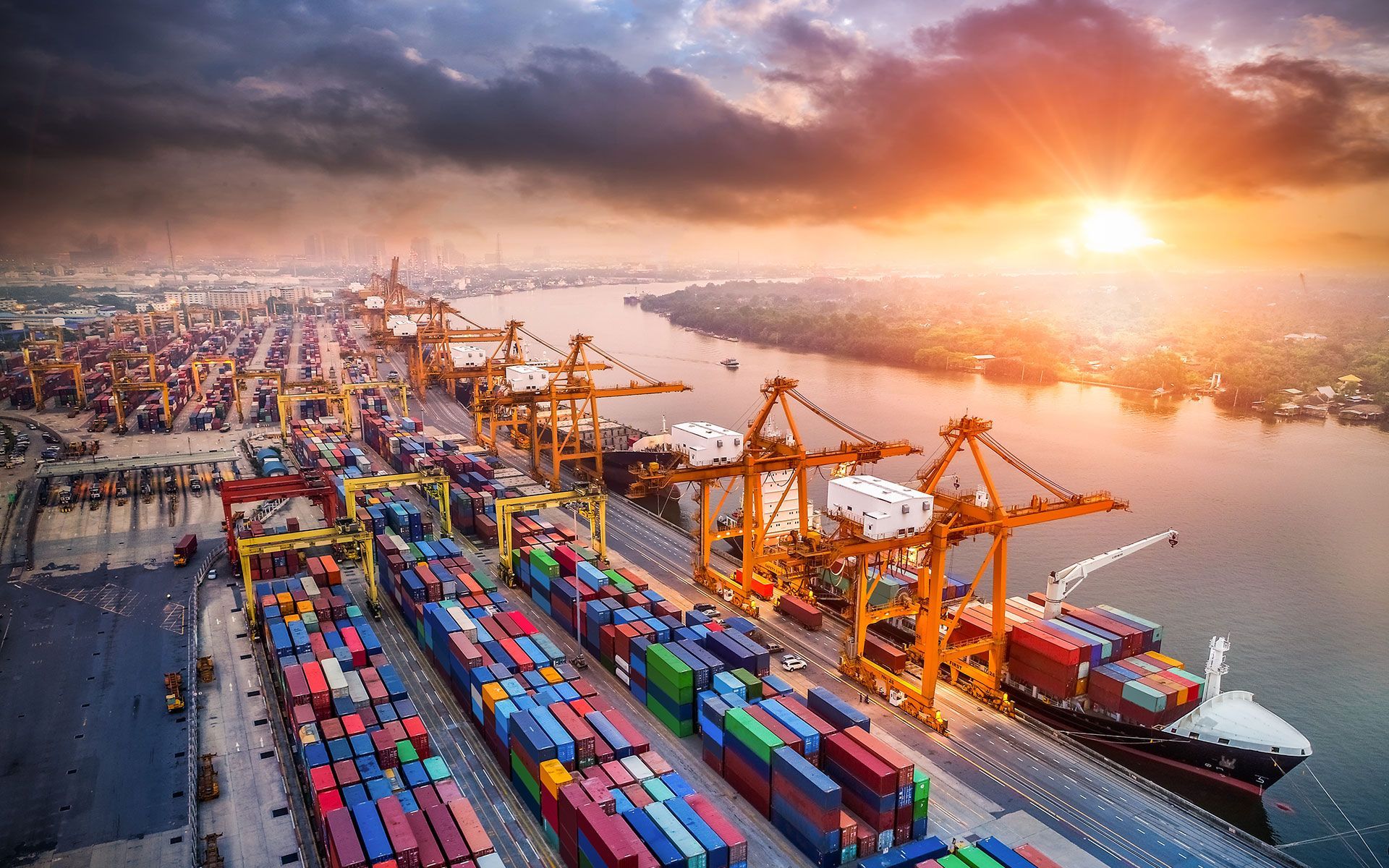 Cargo ships loading containers at a port under a dramatic sunset.