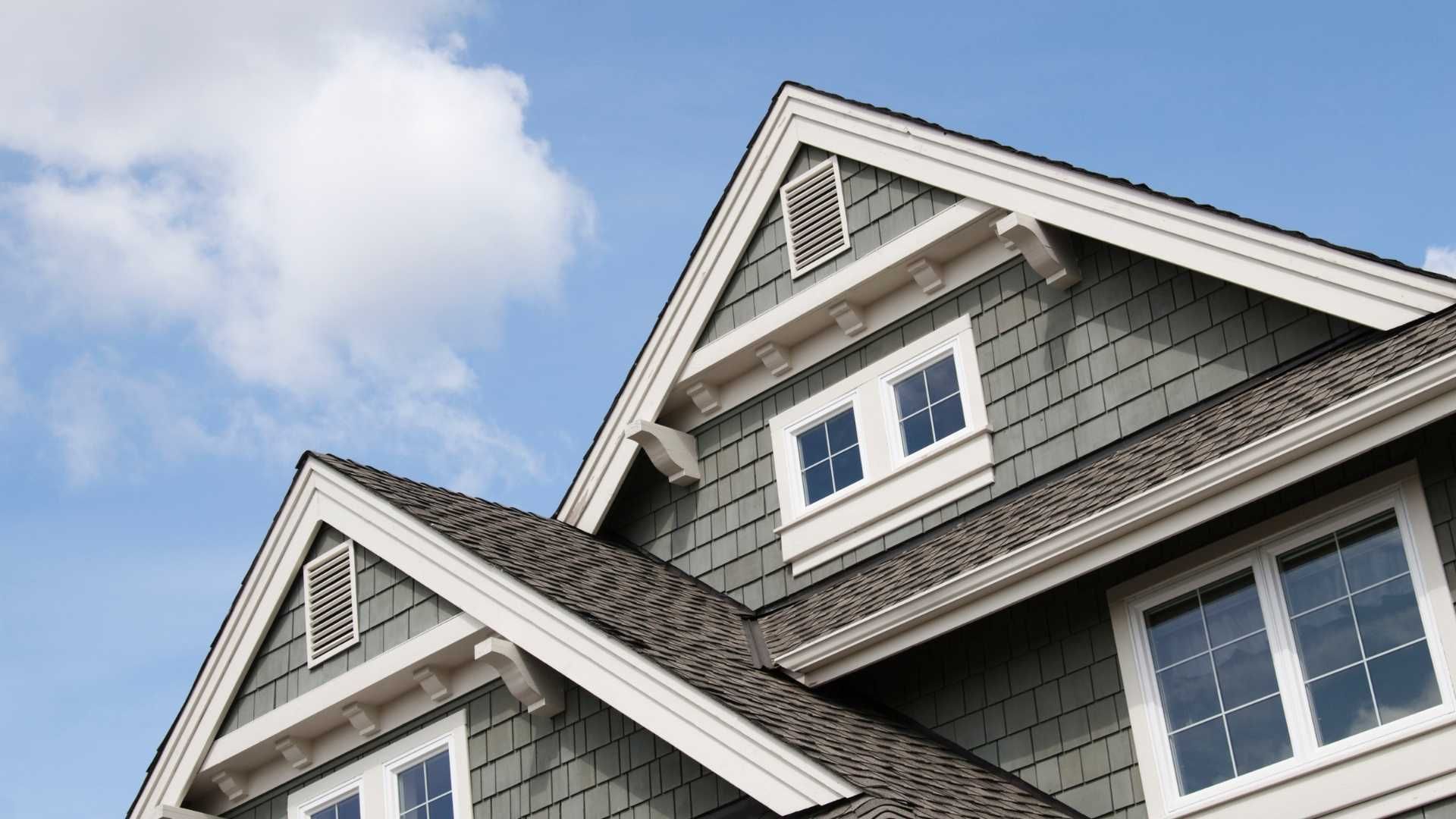 Two gray-shingled house rooftops with white trim against a blue sky