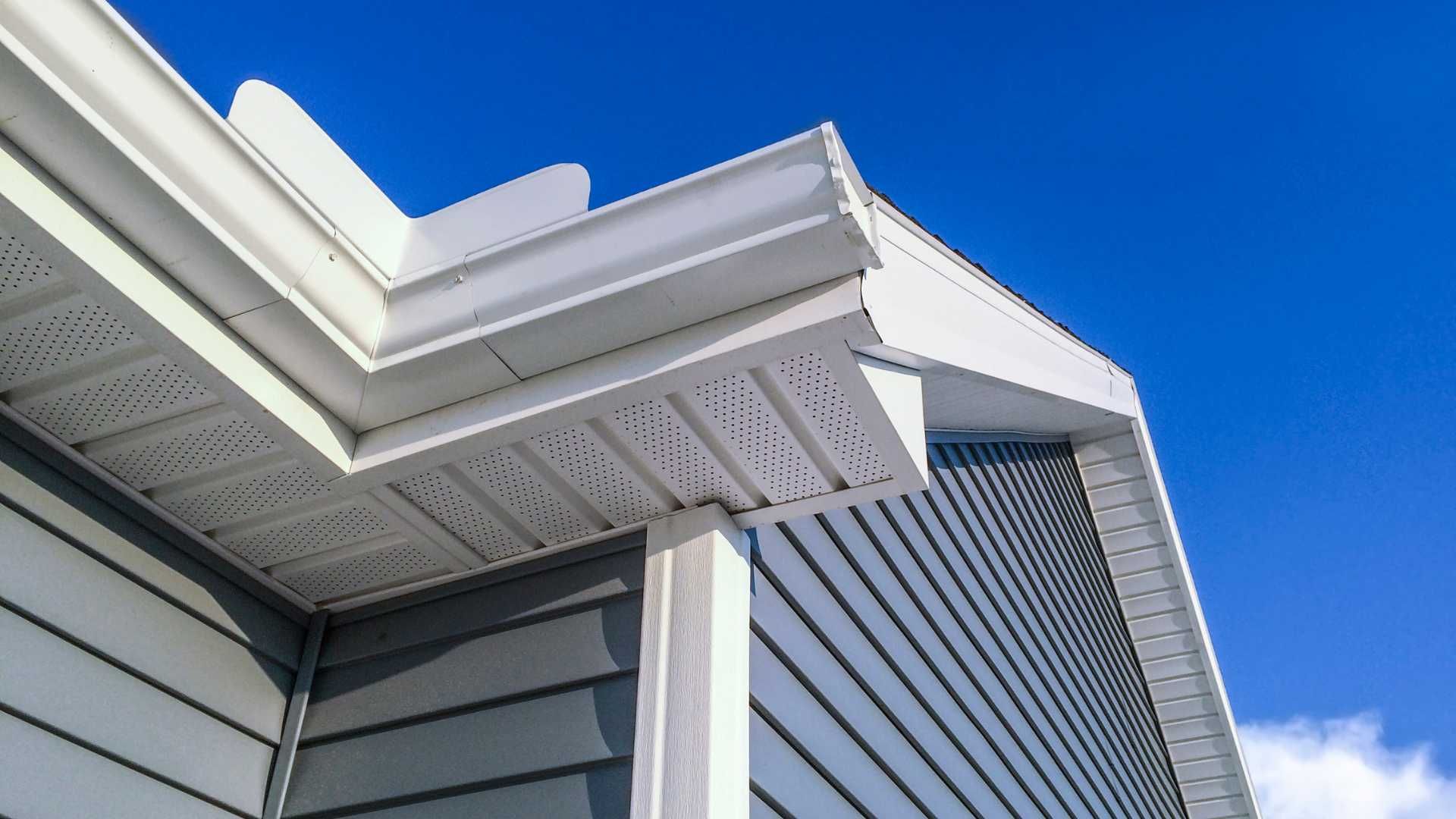 Close-up of a house roof and gable with white trim and gray siding against a blue sky.