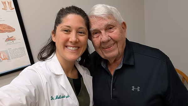 A smiling person in a white coat poses with an elderly person, both in a medical setting.