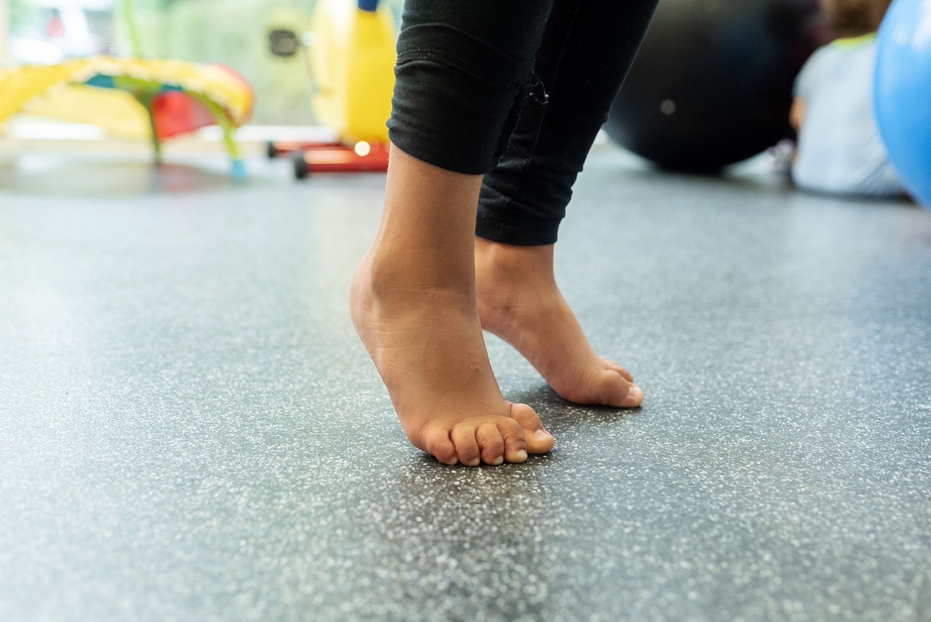 Person's feet on tiptoes on a gray floor, wearing black leggings. Indoors, with exercise equipment in the background.