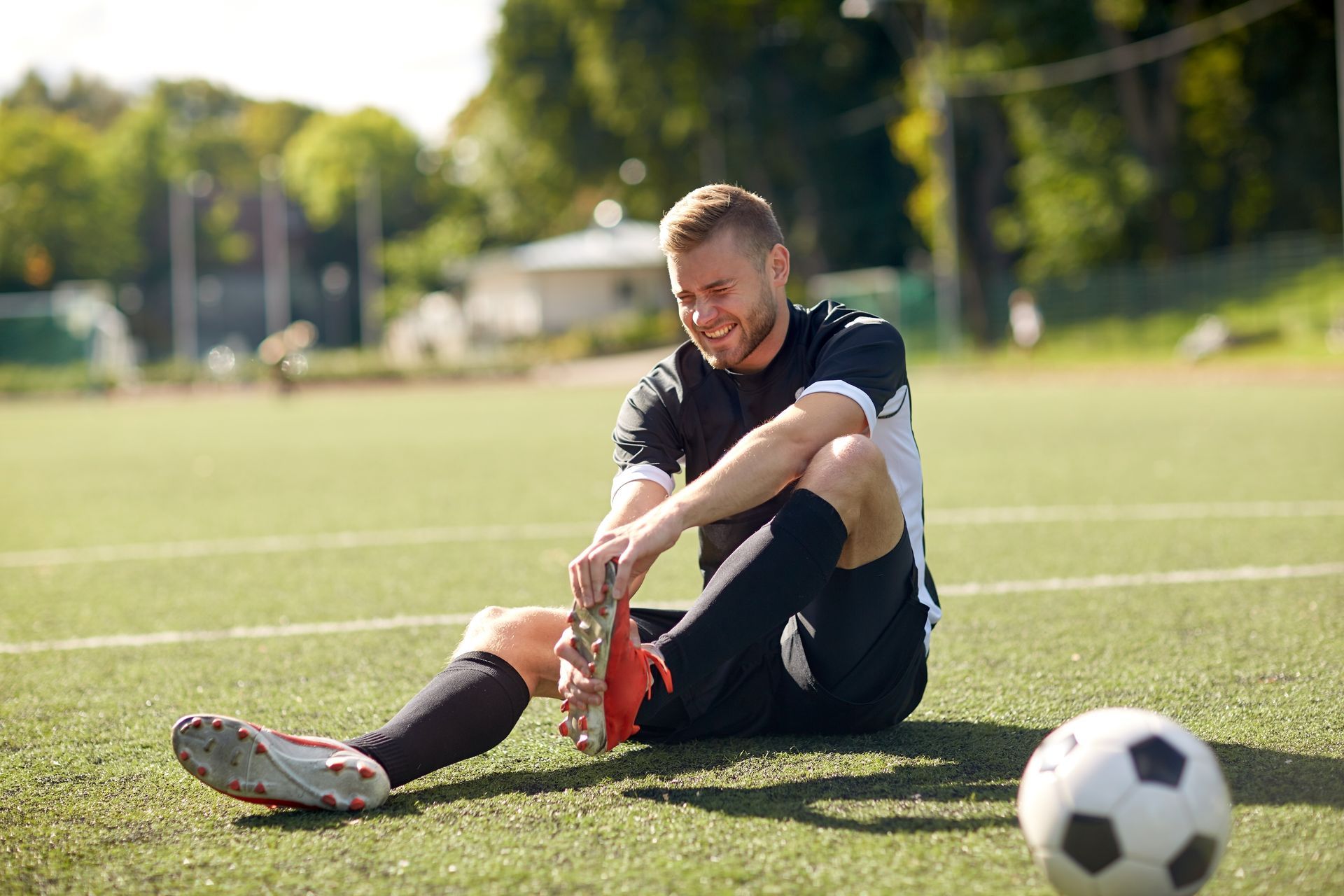 Soccer player with a painful expression holding his ankle, sitting on a green field. A soccer ball is beside him.