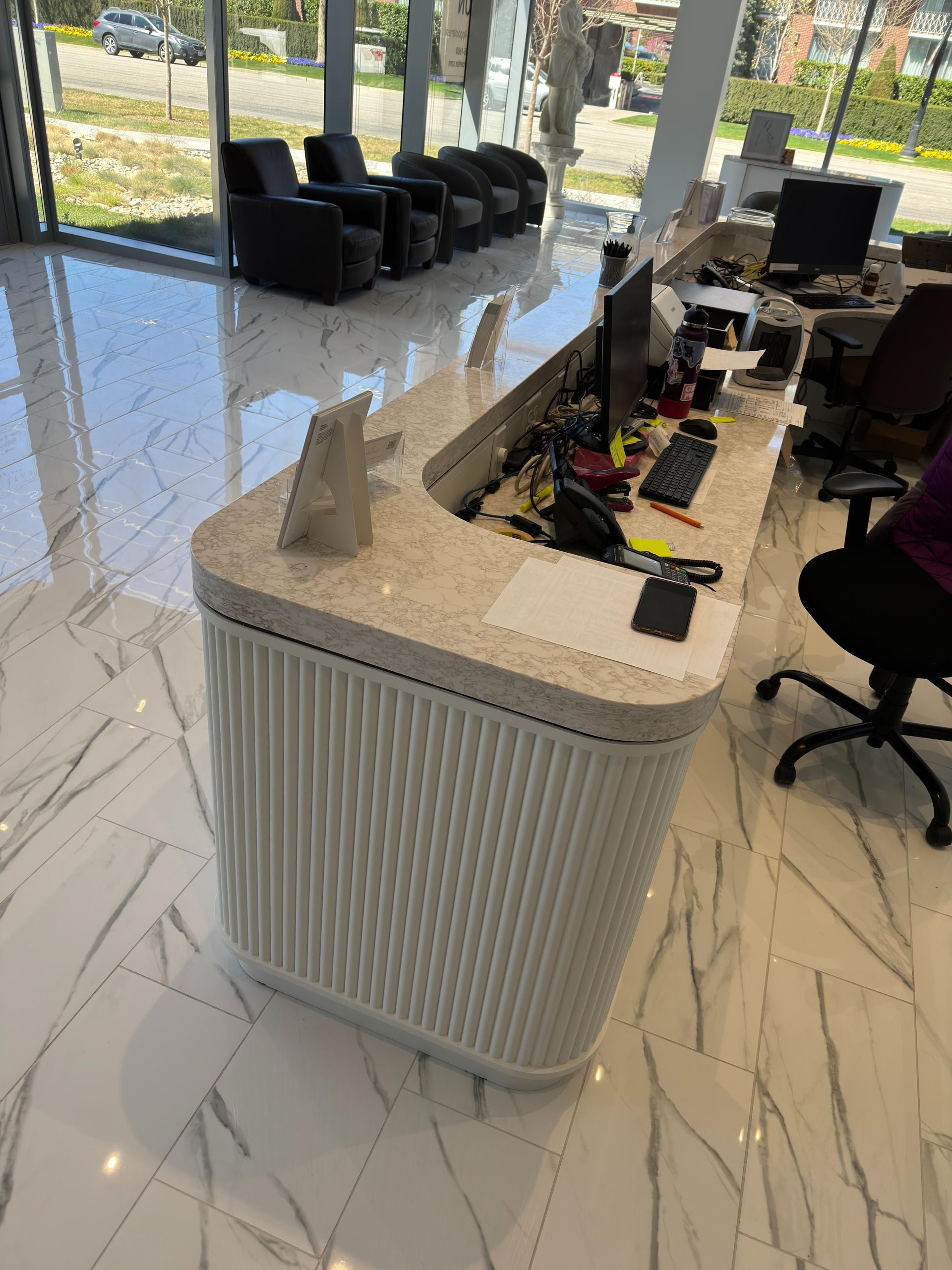 Reception desk with cream-colored countertop and white fluted base. Office setting with seating visible in the background.