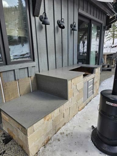 Outdoor kitchen with gray counter, stone base, and black lamps. Snow on the ground.