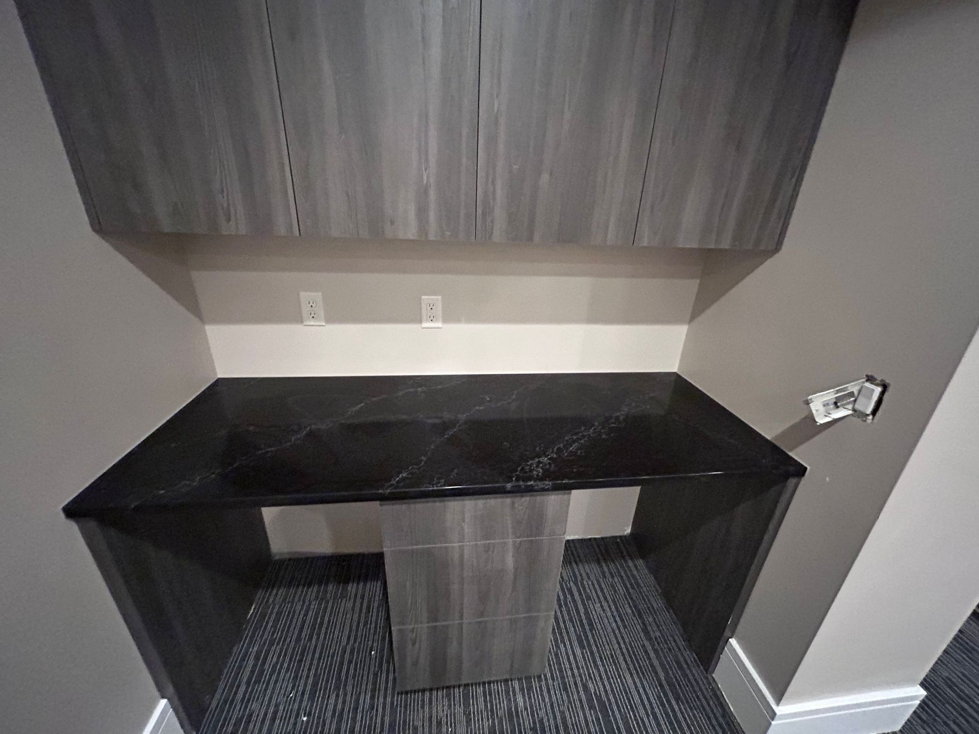 A built-in desk with dark countertop, gray cabinets above, two electrical outlets, and a short central support.