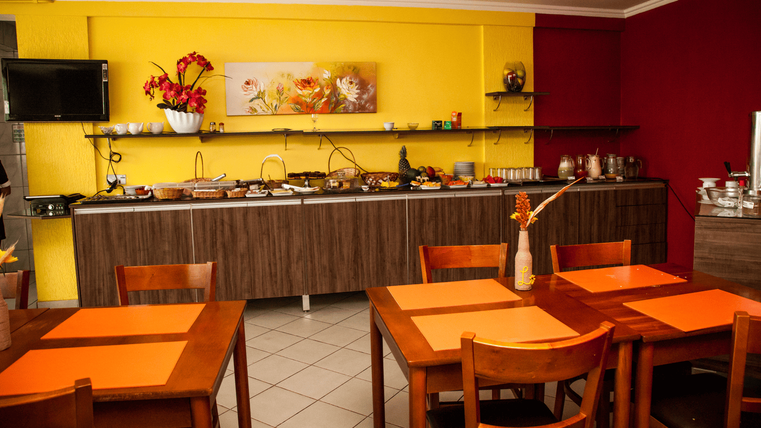 Uma sala de jantar com mesas e cadeiras e uma televisão na parede.