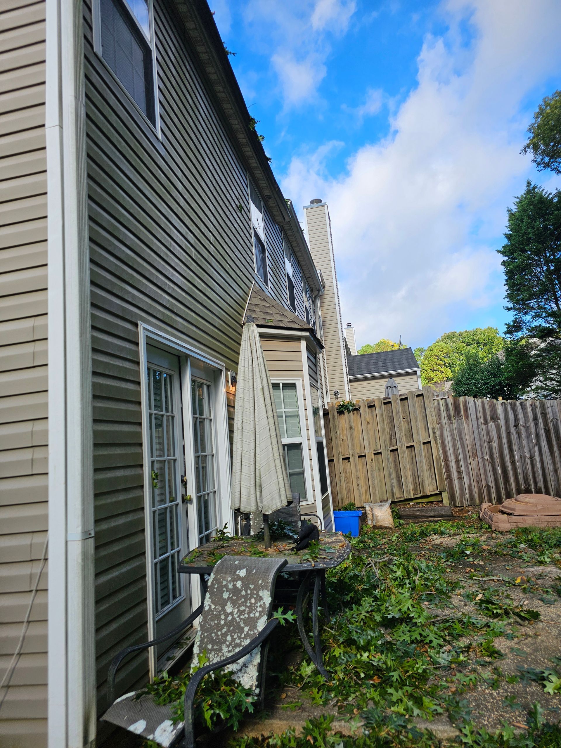 A low-angle view of a beige house exterior with a side patio, debris, and a wooden fence under a blue, cloudy sky.
