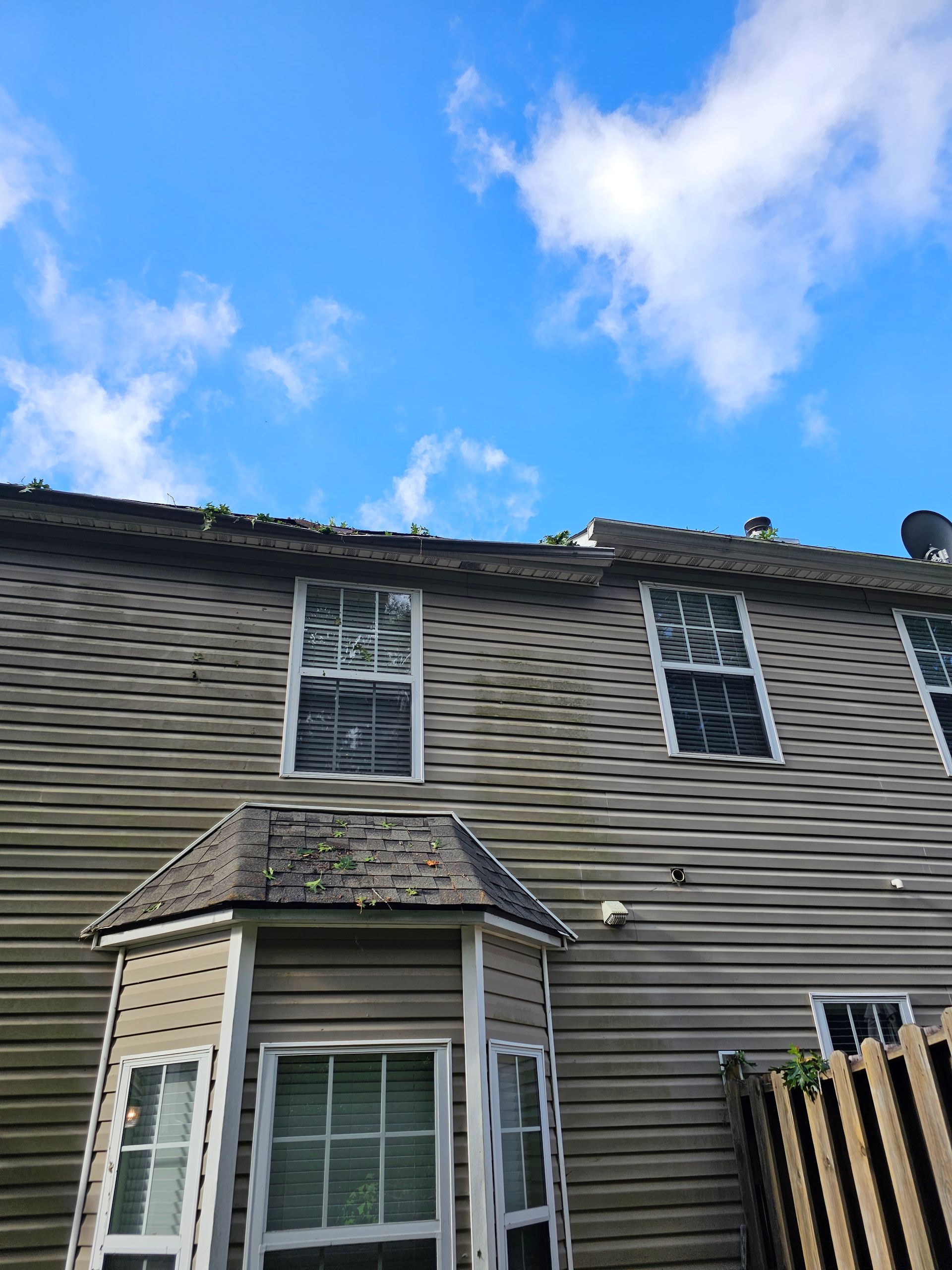 A two-story tan vinyl-sided house with a bay window, showing signs of dirt and moss under a blue sky with white clouds.