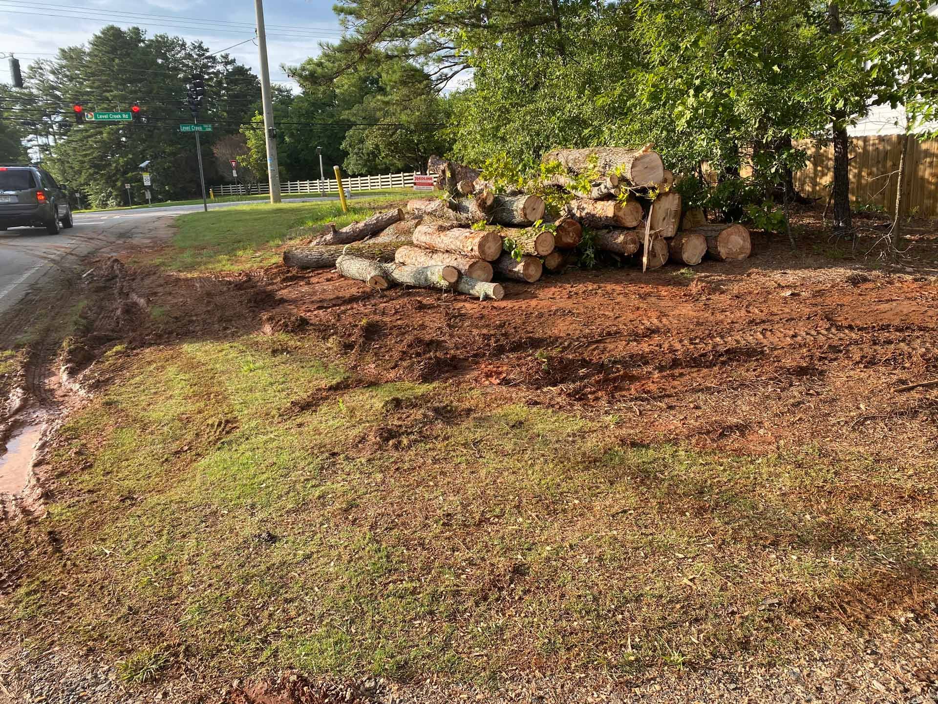 A pile of stacked tree logs rests on cleared red earth beside a grassy roadside and a passing vehicle.