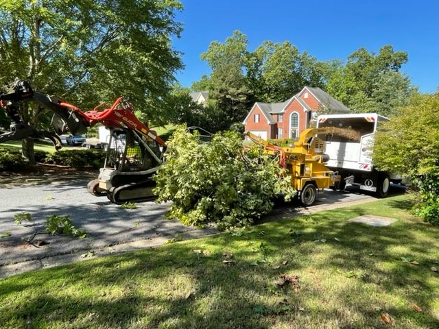 A red skid steer and yellow wood chipper process tree branches in a residential driveway near a brick house.
