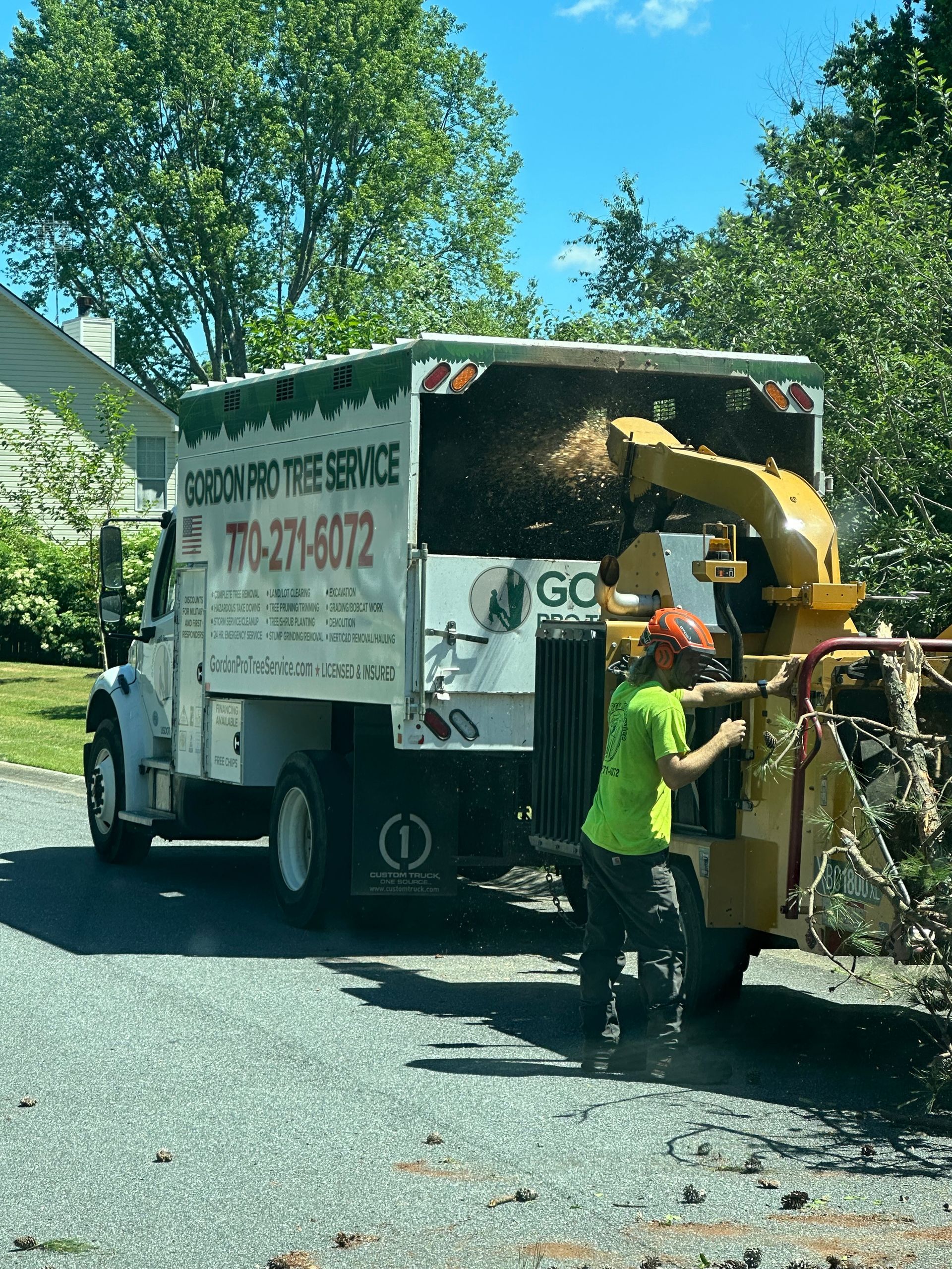 A worker in a neon green shirt feeds tree branches into a wood chipper attached to a white truck on a gravel driveway.