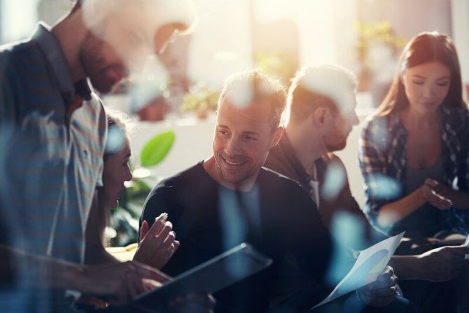 Group of people in a meeting, smiling and looking at documents in a brightly lit room.