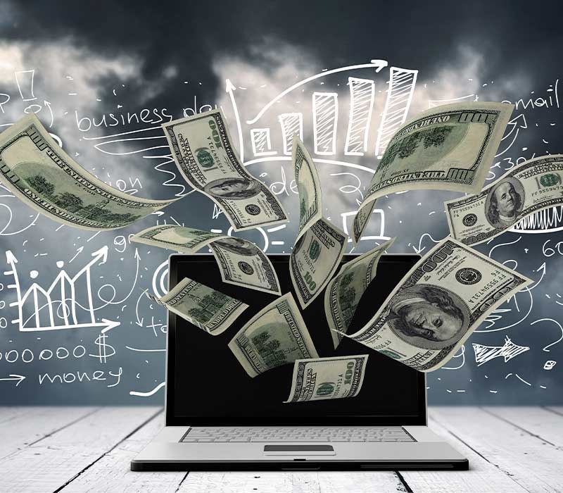 Laptop with money flying out; business drawings in background.