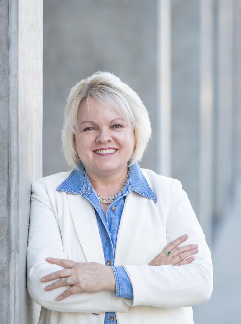 Woman in white blazer, denim shirt, smiling, arms crossed, leaning against concrete pillars.
