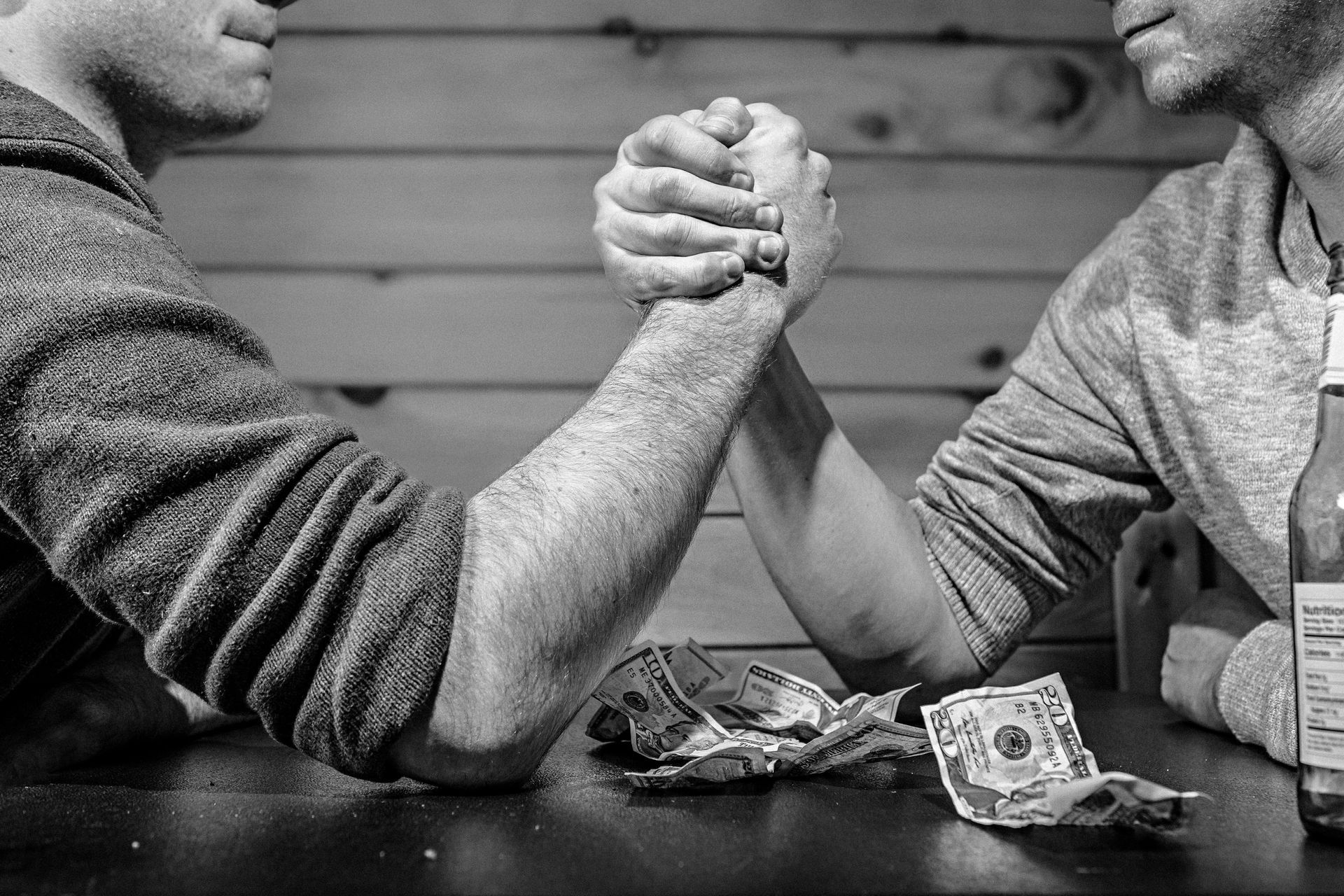 Two men arm wrestling, money on table, dark, indoor setting.