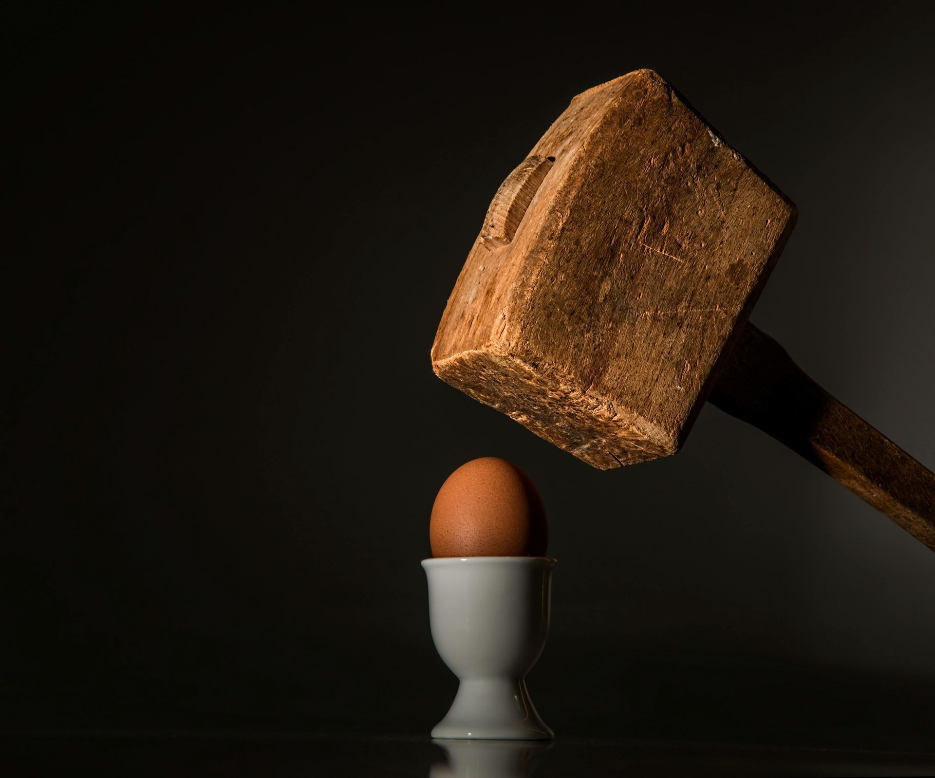 Wooden mallet about to smash a brown egg in a white egg cup, dark background.