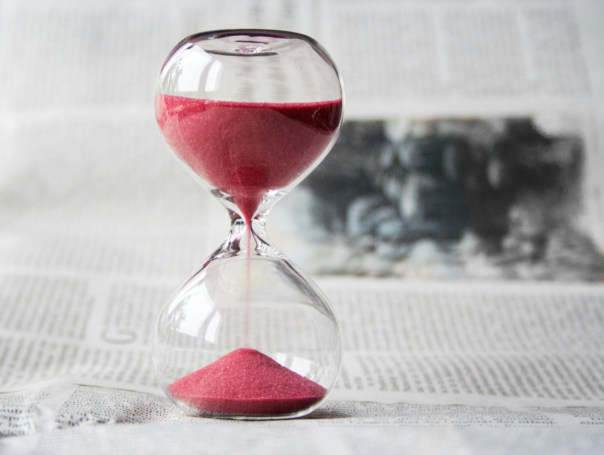 Hourglass with red sand, upper chamber nearly empty, lower chamber full. Set against a newspaper background.