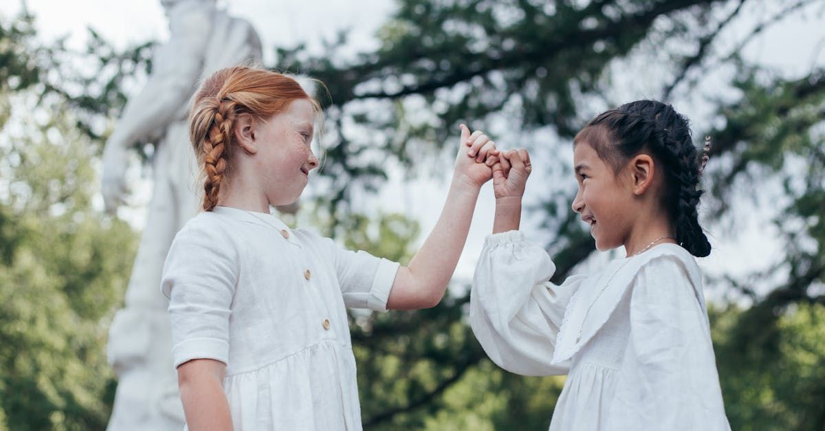 Two girls in white dresses touch pinky fingers, smiling, near a statue outdoors.