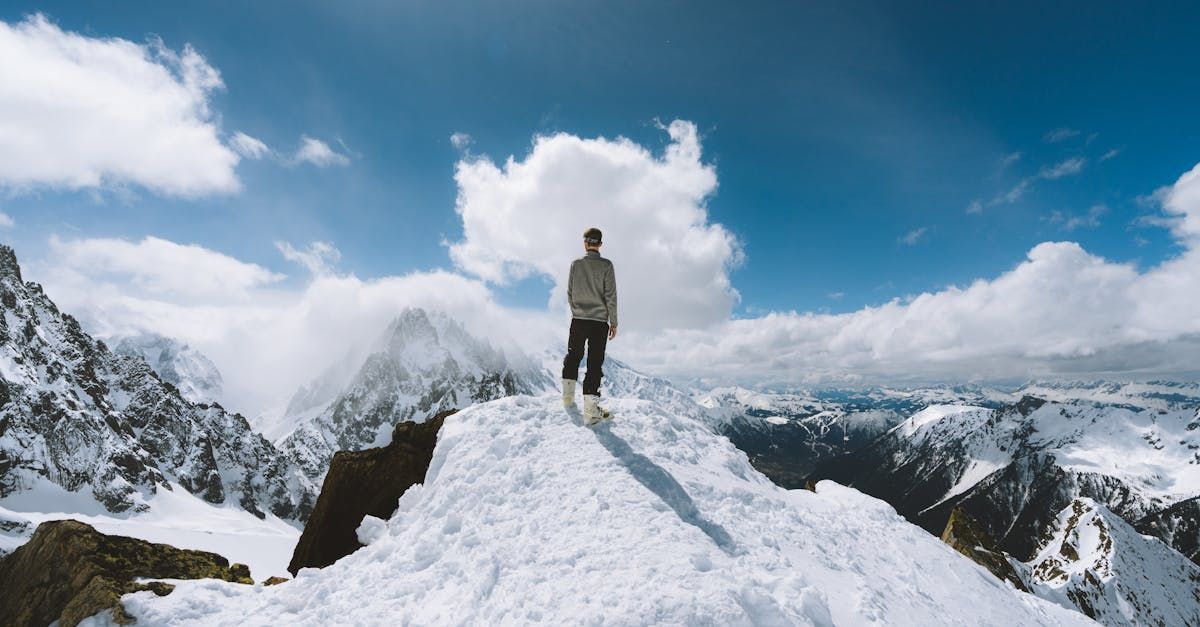 Person stands on snowy mountain peak, looking at distant snowy mountains under blue sky with clouds.