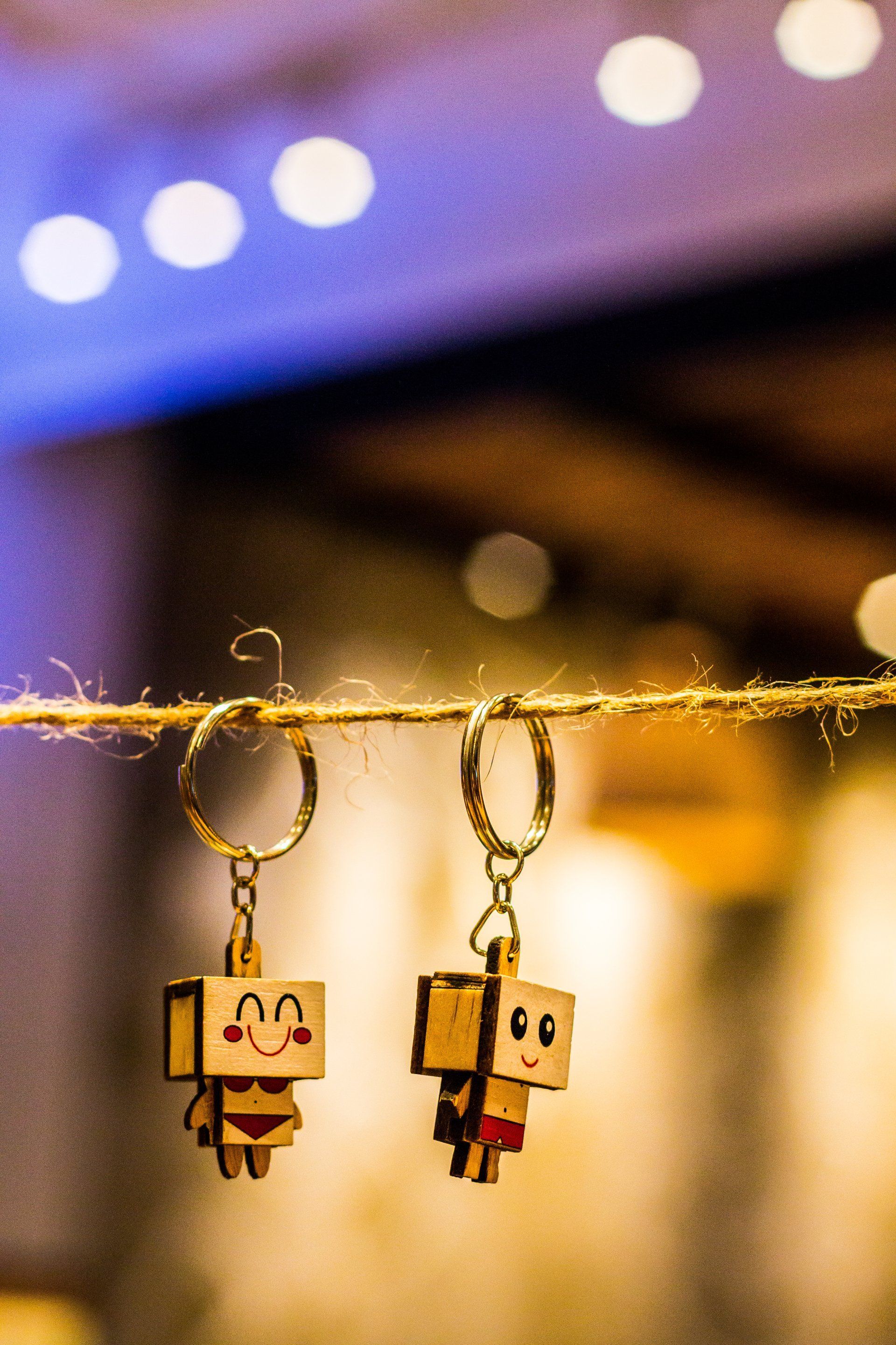 Two wooden keychains with cartoon-like figures, hanging on a string, against a blurry background.
