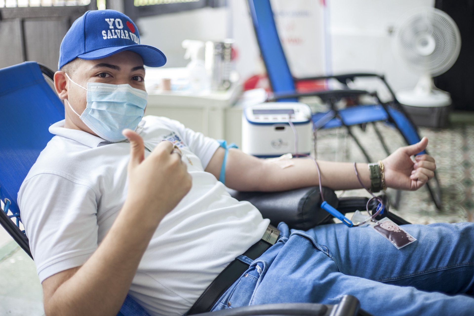 Man donating blood, wearing a mask and blue hat, giving a thumbs up in a medical setting.