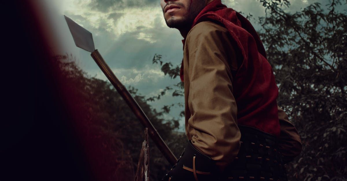 Person in a red hood, holding a spear, looking up towards a cloudy sky.
