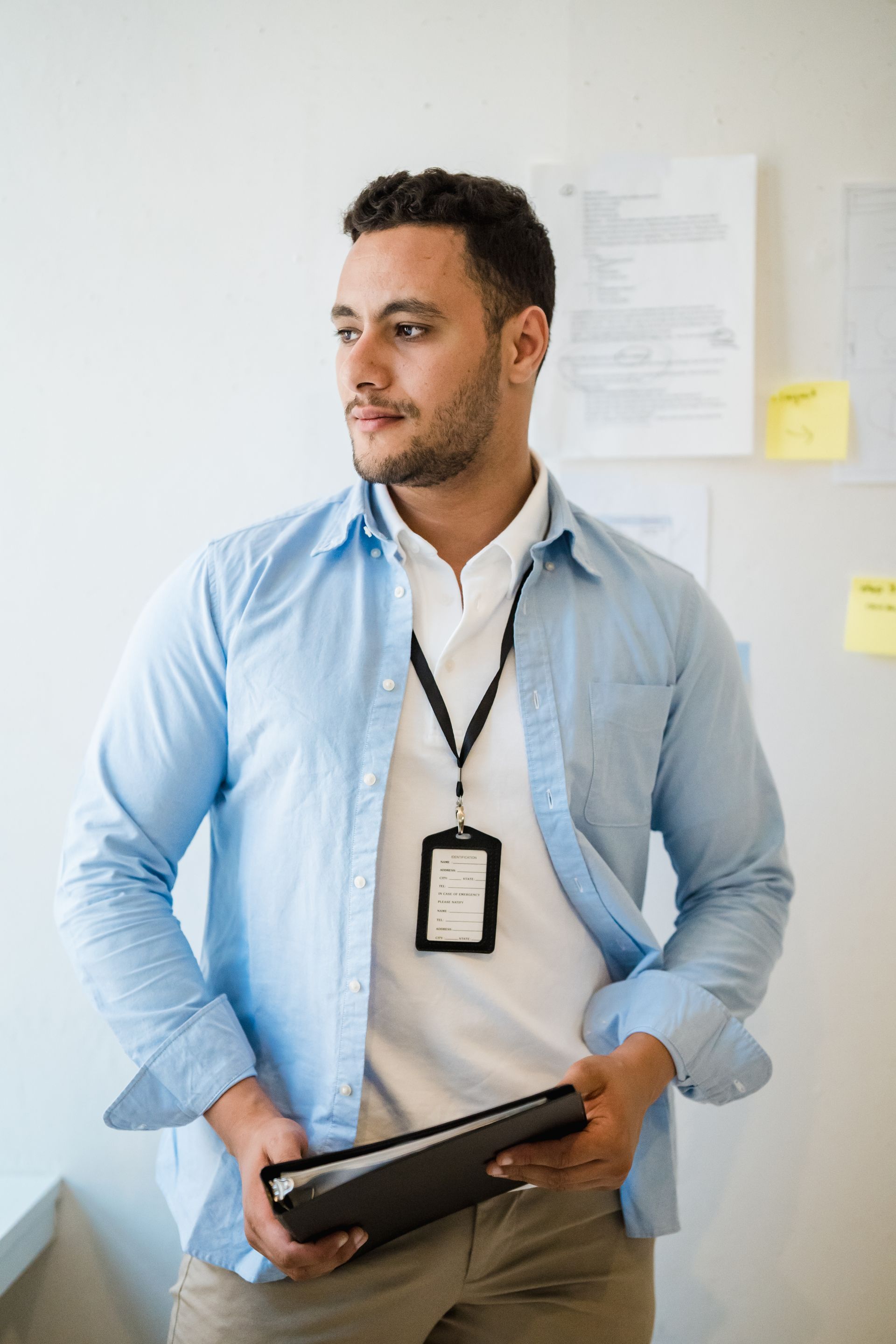 Man with lanyard and papers, wearing open blue shirt, looking away, near a white wall with papers.