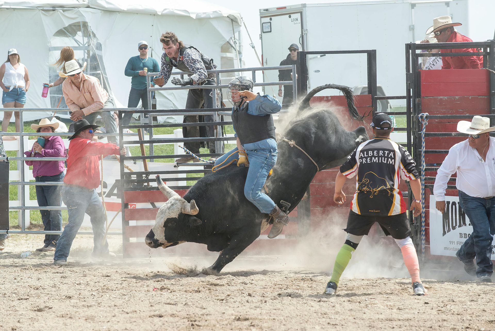 Bull rider on black bull in rodeo arena, surrounded by spectators and protective personnel.