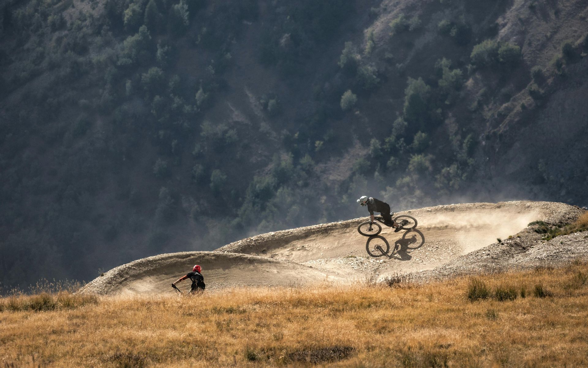 Two mountain bikers on a dirt track, one airborne, in a mountainous, dry landscape.