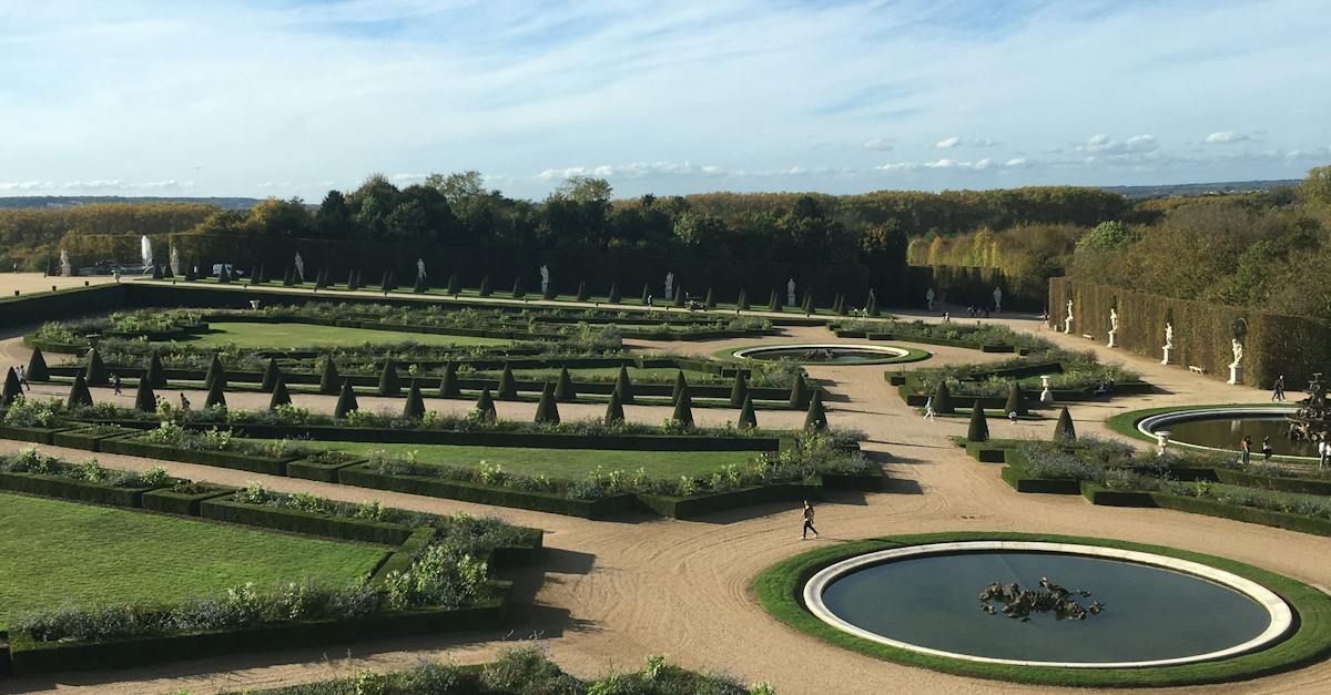 Formal garden with symmetrical paths, fountains, and greenery under a blue sky.