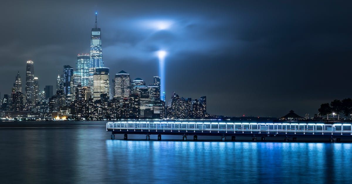New York City skyline at night with two beams of light rising. Blue water and a pier in foreground.