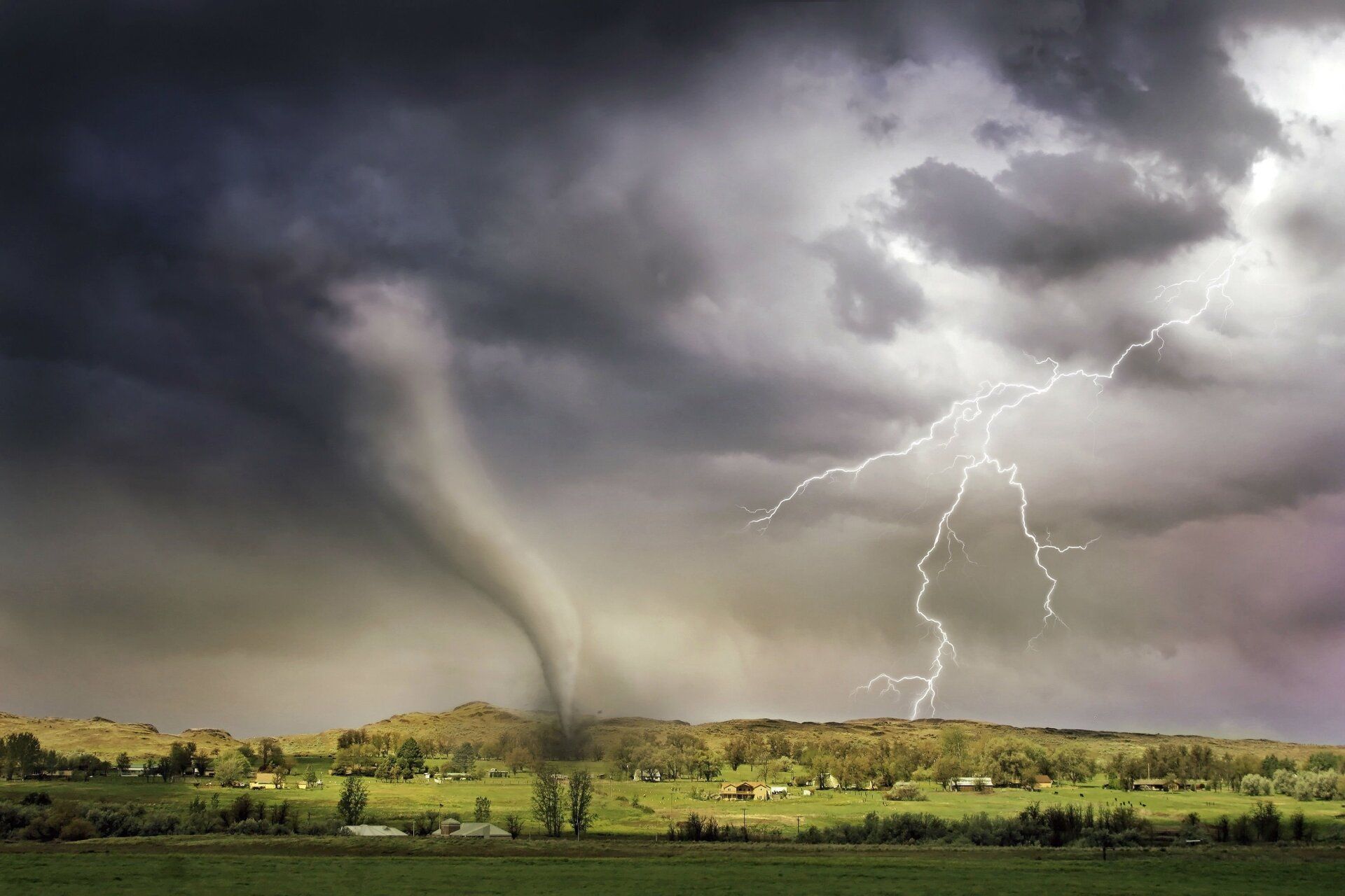 Tornado descends over a green landscape with lightning striking in a stormy sky.