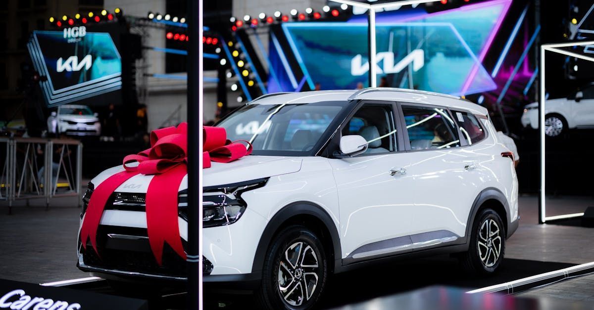 White Kia Carens SUV with red bow at an event, stage with Kia logo backdrop.