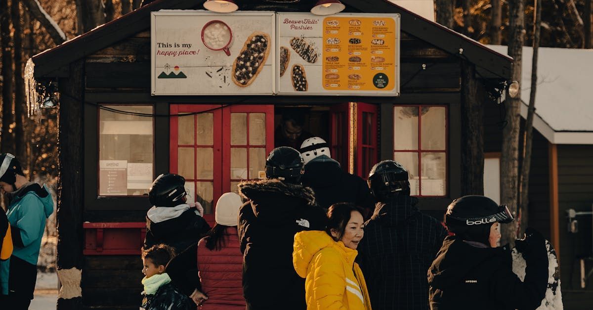 People lined up at a winter food stand with menu signs. Trees and snow visible.