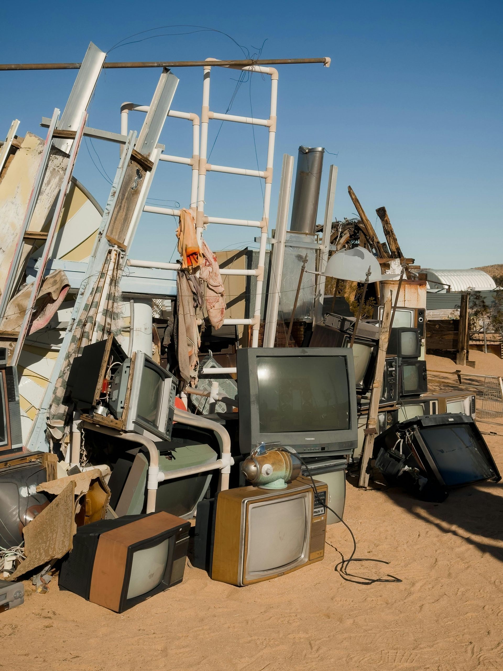 Pile of discarded TVs and metal scraps in a desert setting, with a ladder and blue sky.
