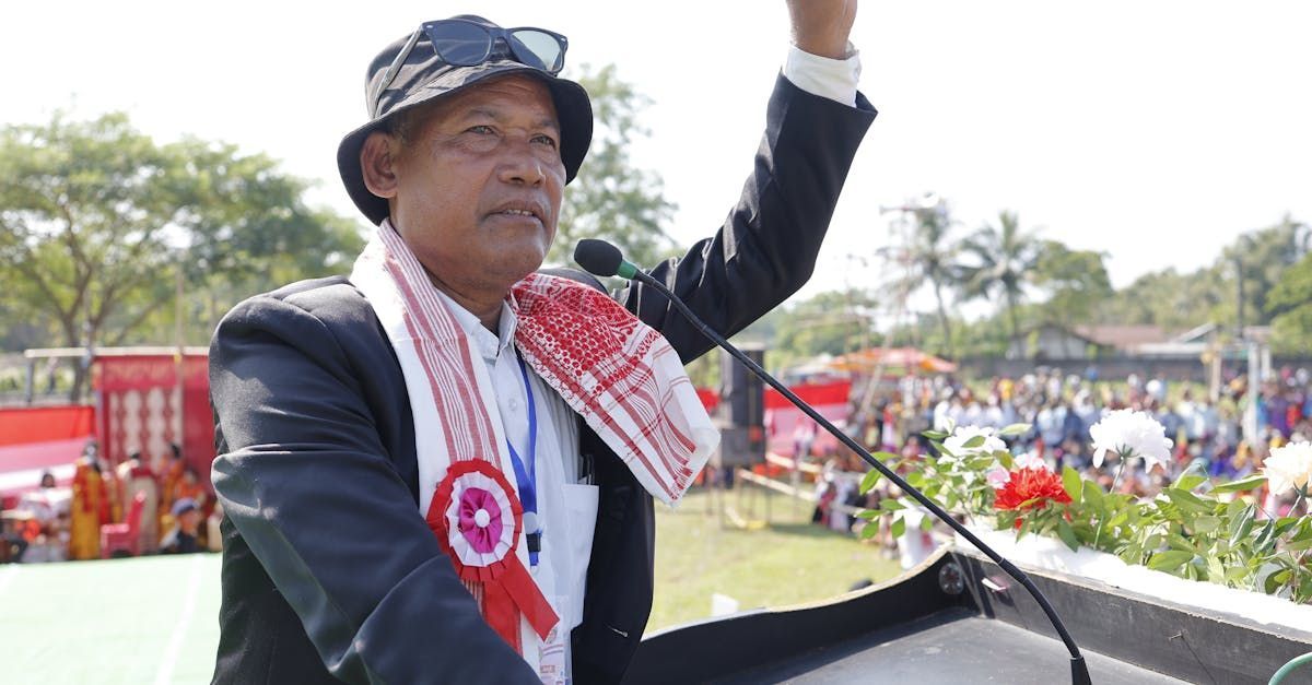 Man speaking at an outdoor event, wearing a suit, hat, and scarf. He is raising his arm.