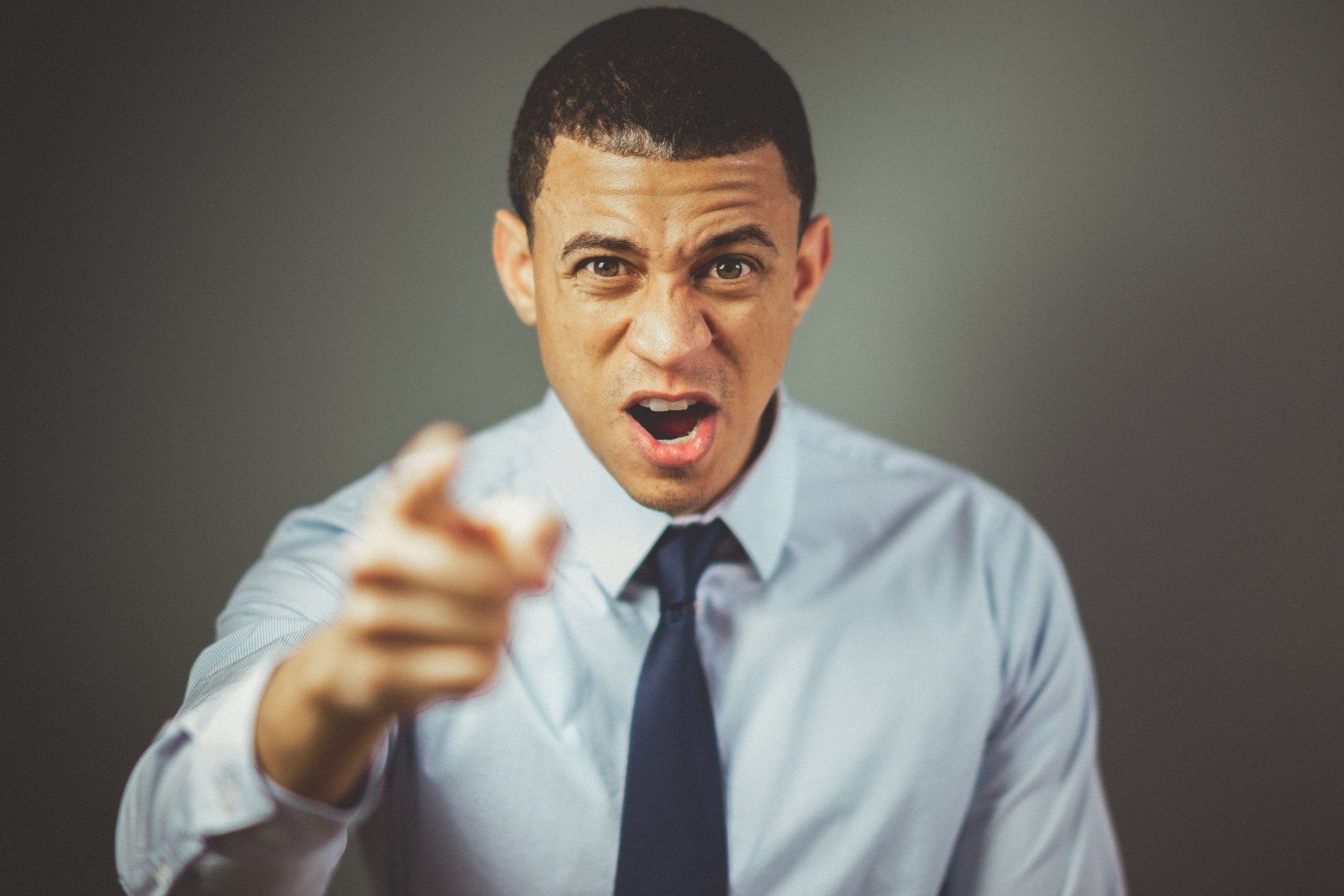 Man in white shirt and tie, pointing with an angry expression, mouth open, against a grey background.