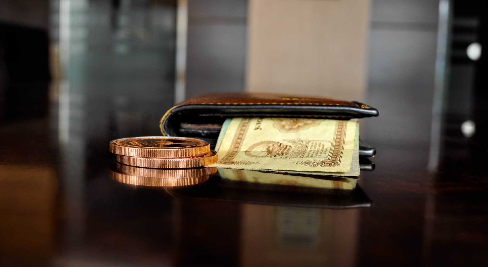 Brown wallet with cash and a stack of copper coins on a reflective surface.