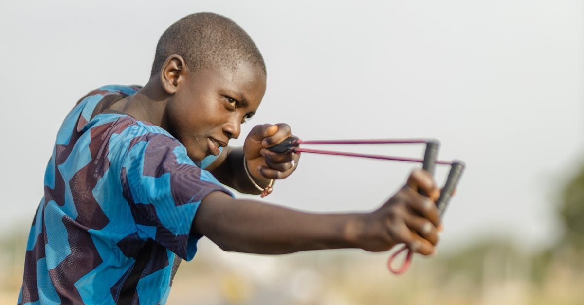 Boy with slingshot, aiming with focused expression, wearing blue patterned shirt.