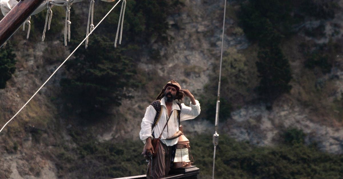 Bearded man in white shirt on ship, looking up, hand on brow, against cliffside.