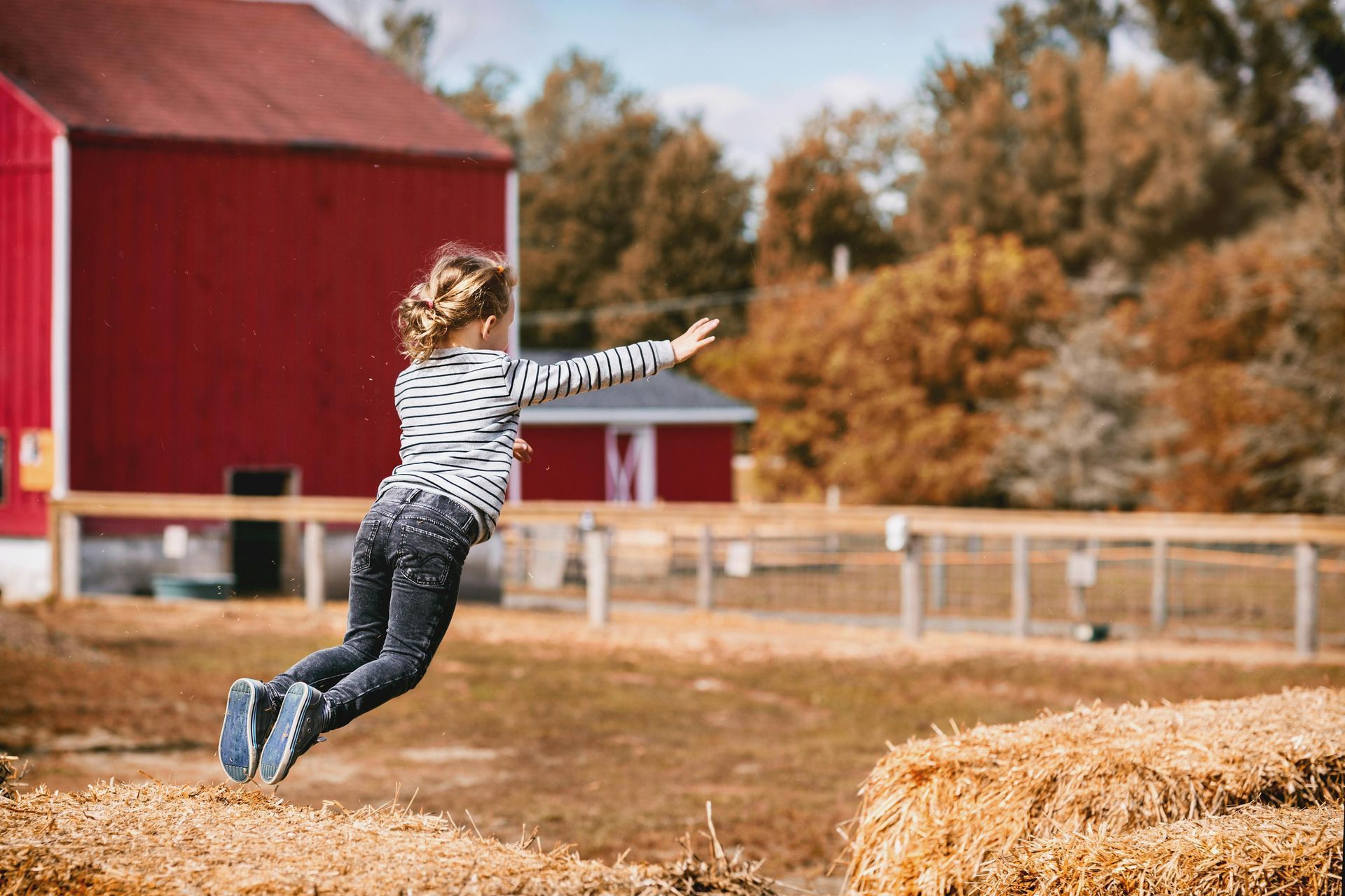 Girl jumping over hay bales near a red barn on a sunny day.