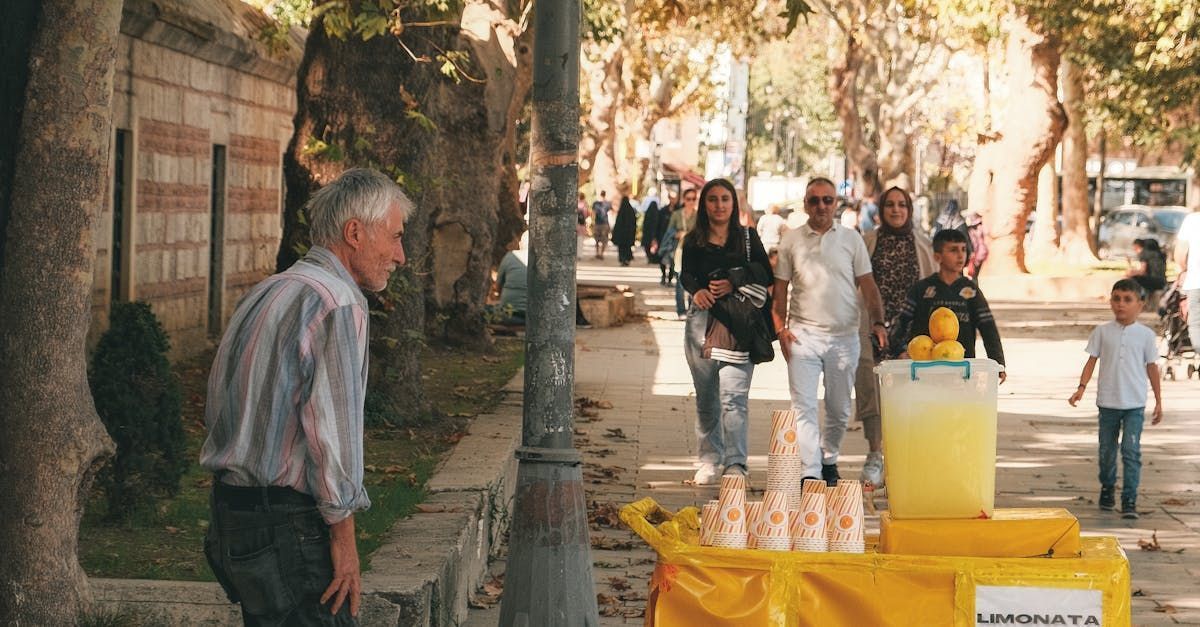 Man near street vendor with lemonade on sunny sidewalk, other people walking.